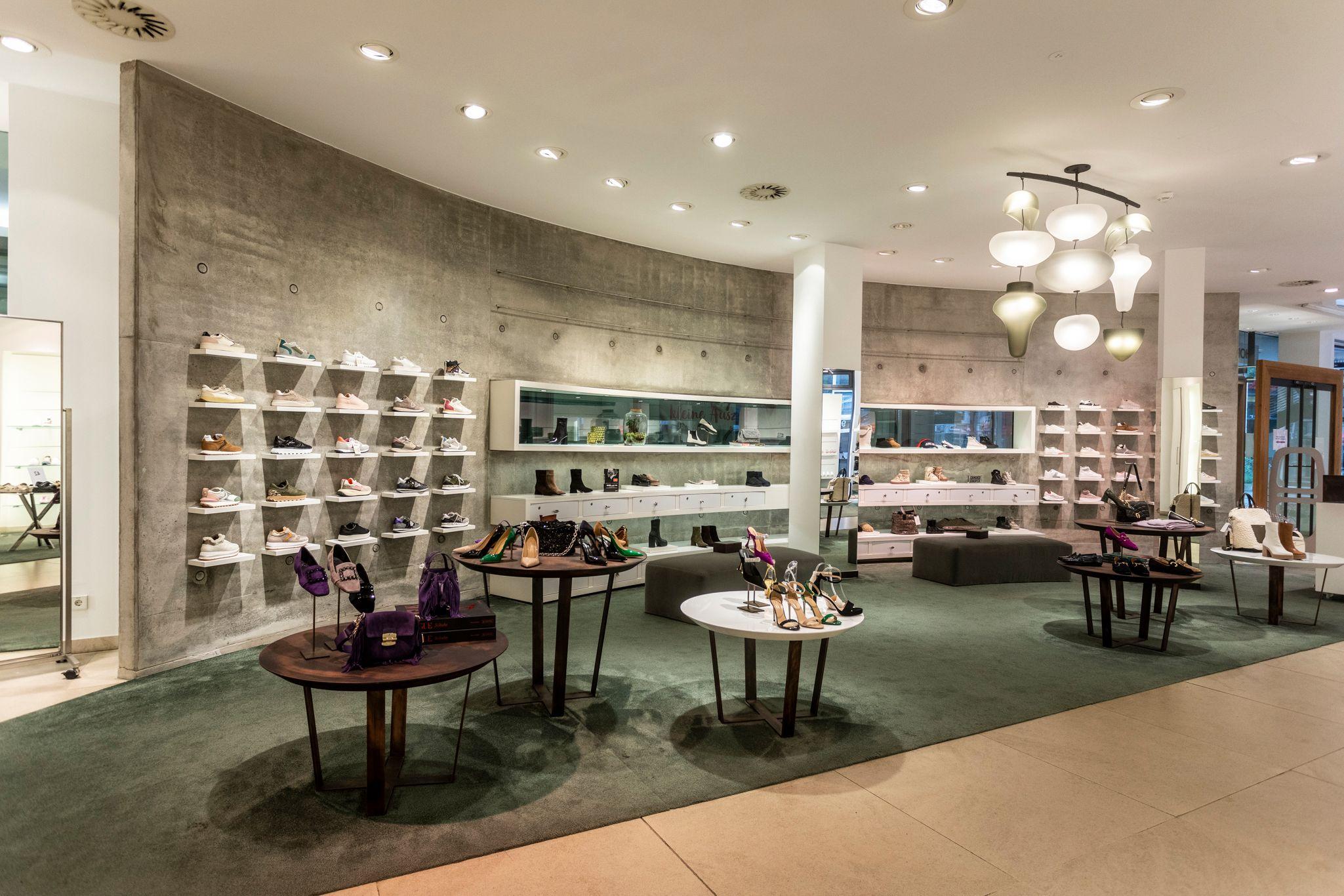 An upscale shoe department featuring shoes on wall shelves, round tables, and a unique chandelier.