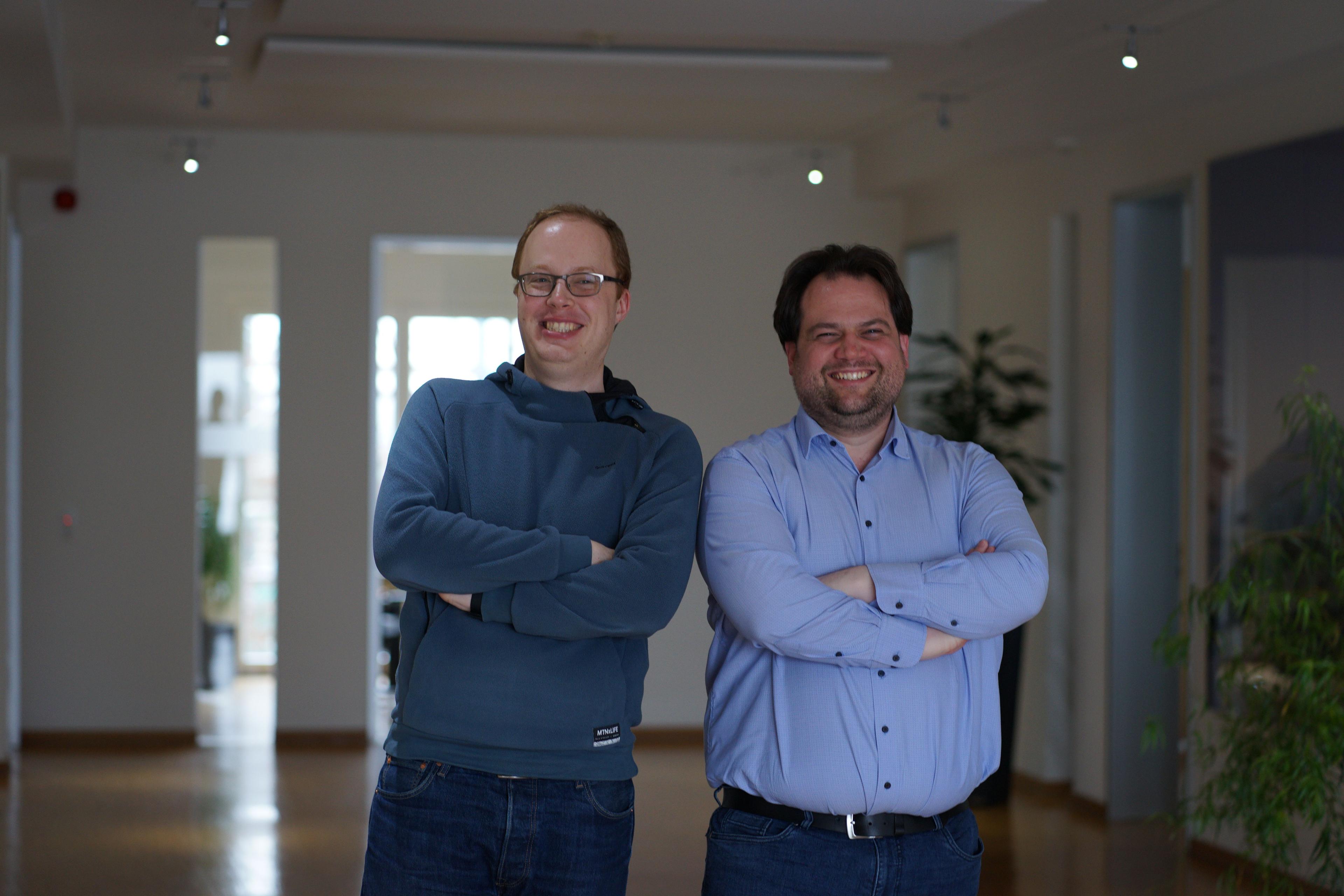 Two smiling men, arms crossed, standing comfortably in a modern office environment.
