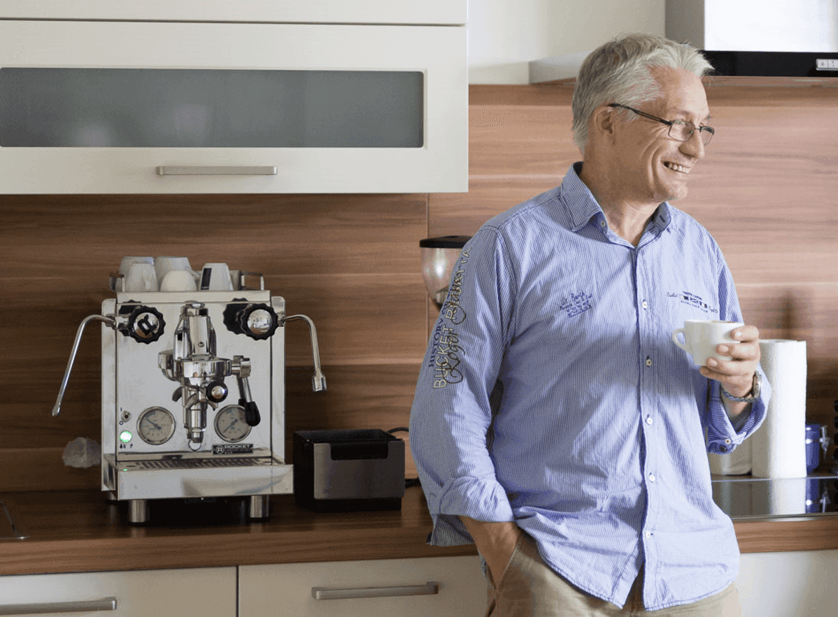 Smiling man with glasses holds a coffee cup near an espresso machine in a modern kitchen.