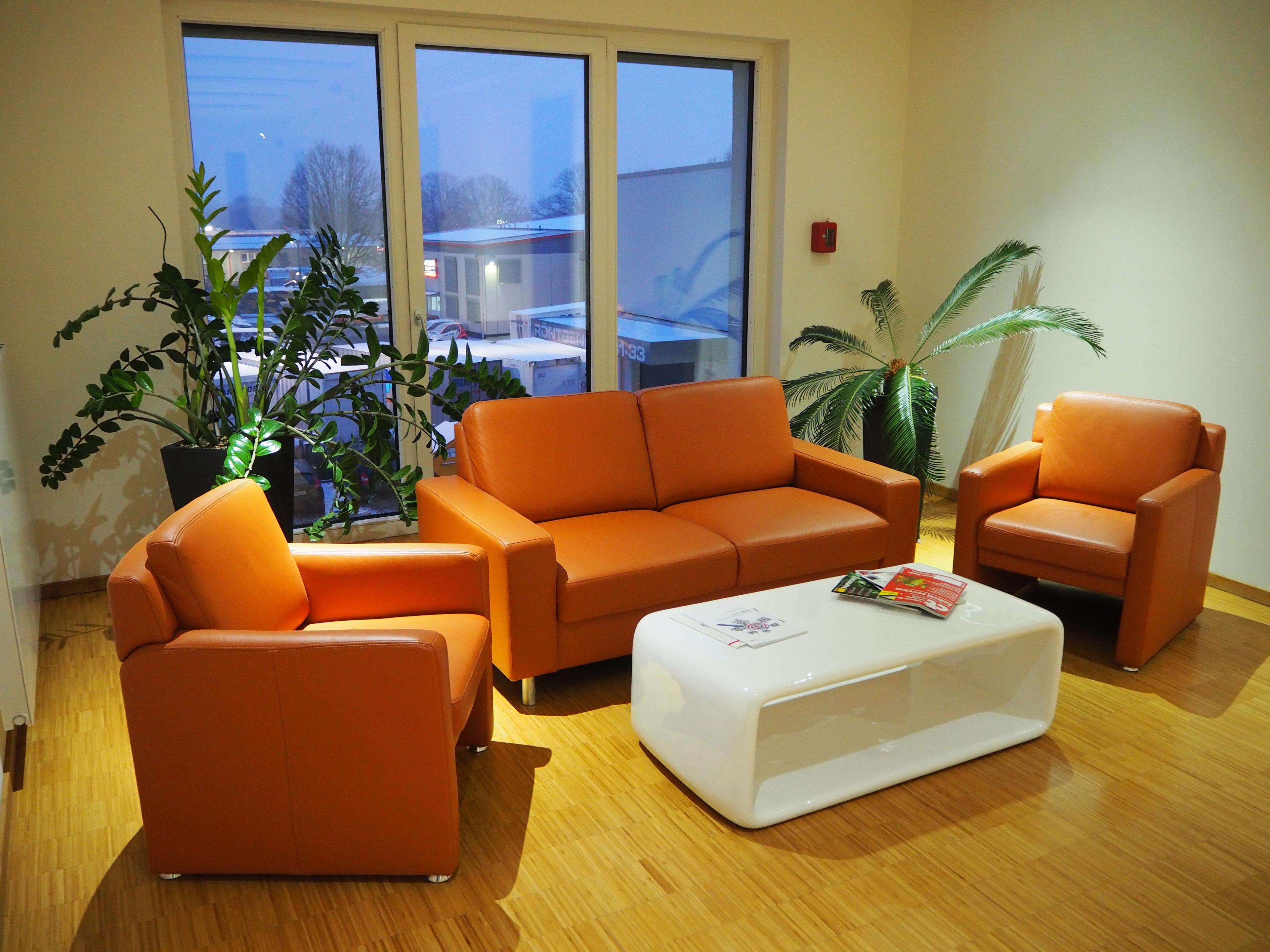 Modern waiting room with orange sofa, armchairs, white table, and green potted plants by a window.
