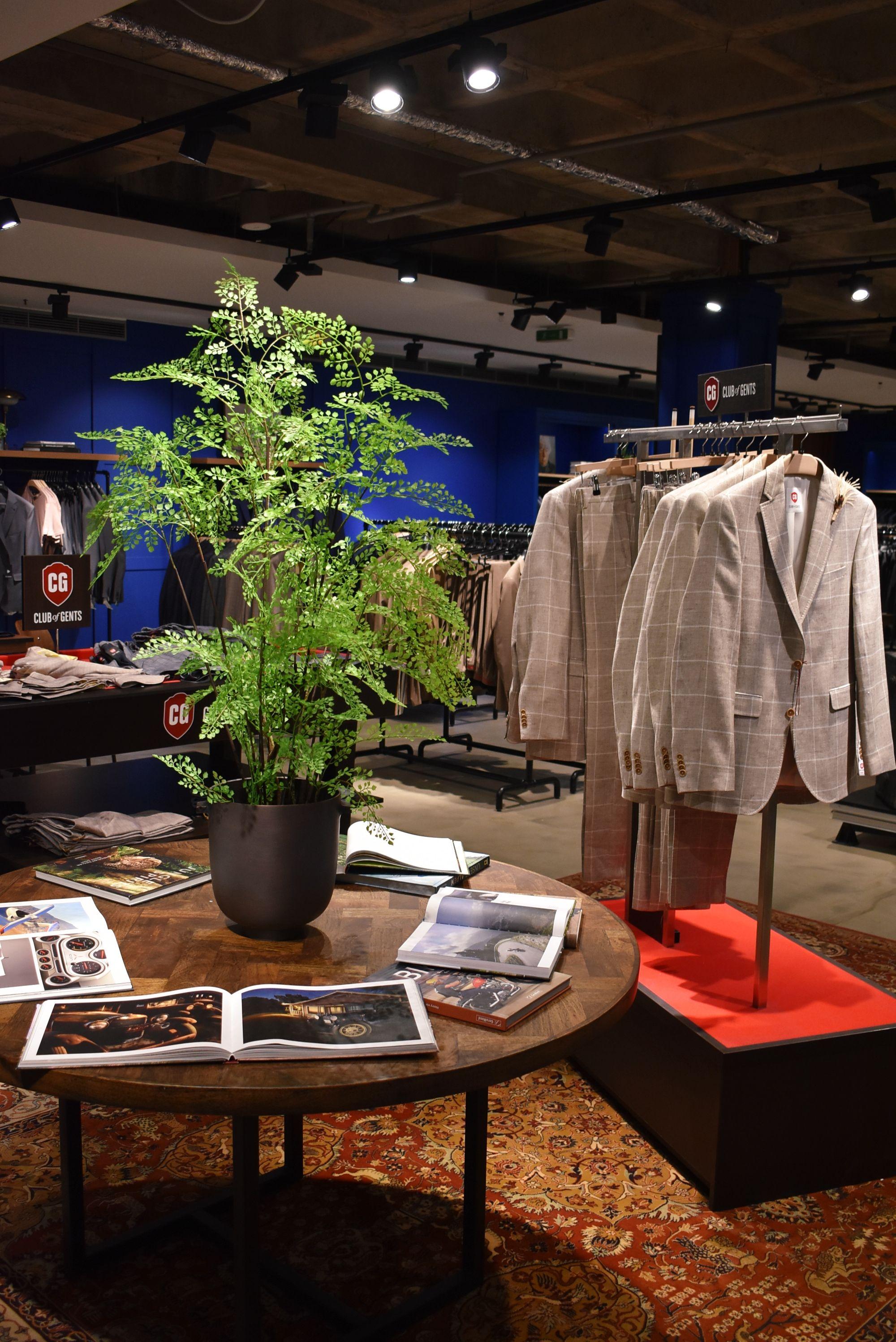 Store interior featuring a round wooden table with a plant, magazines, and light blazers.