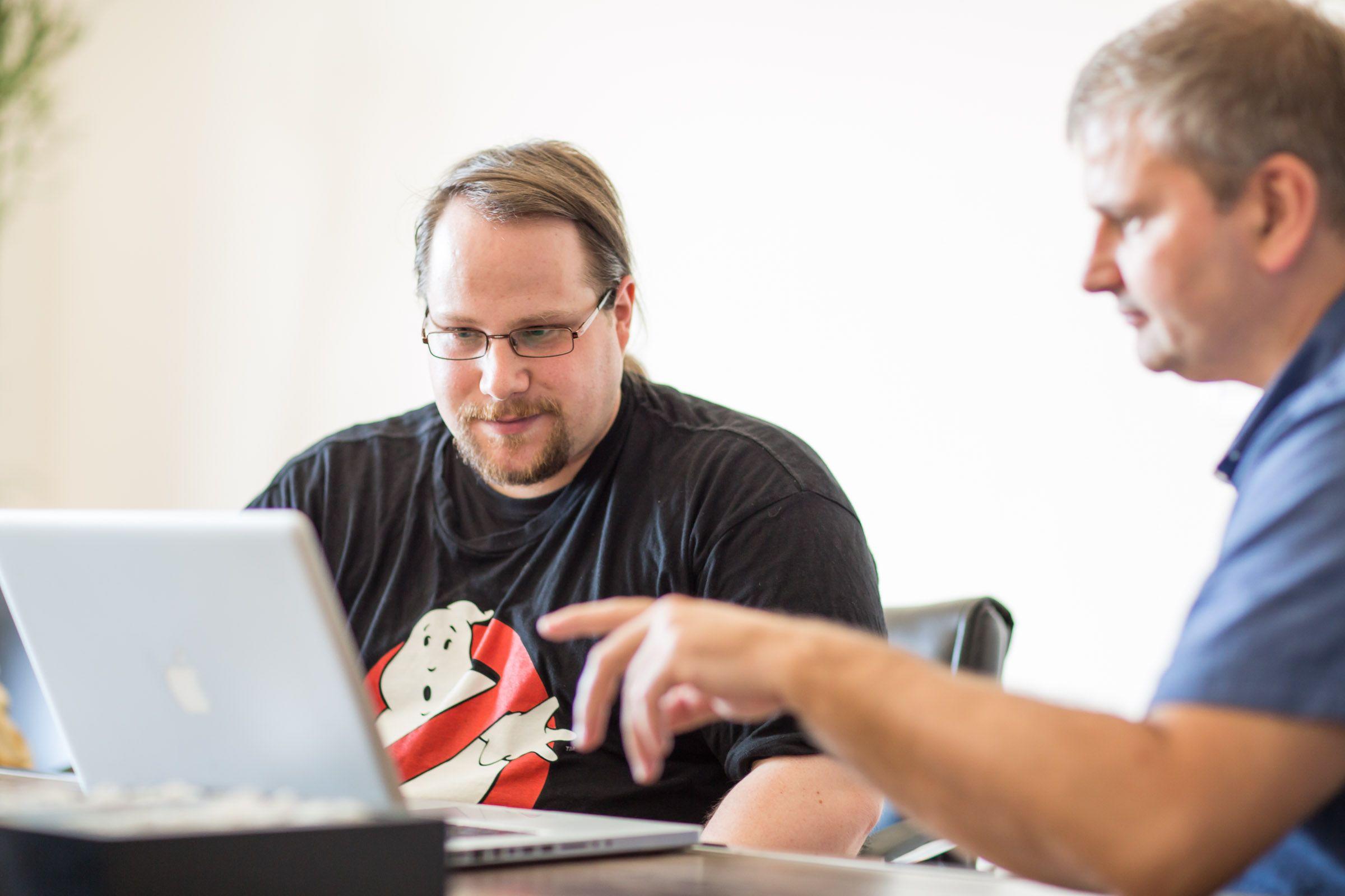 Two men collaborate on a laptop, one wearing a Ghostbusters t-shirt and glasses.