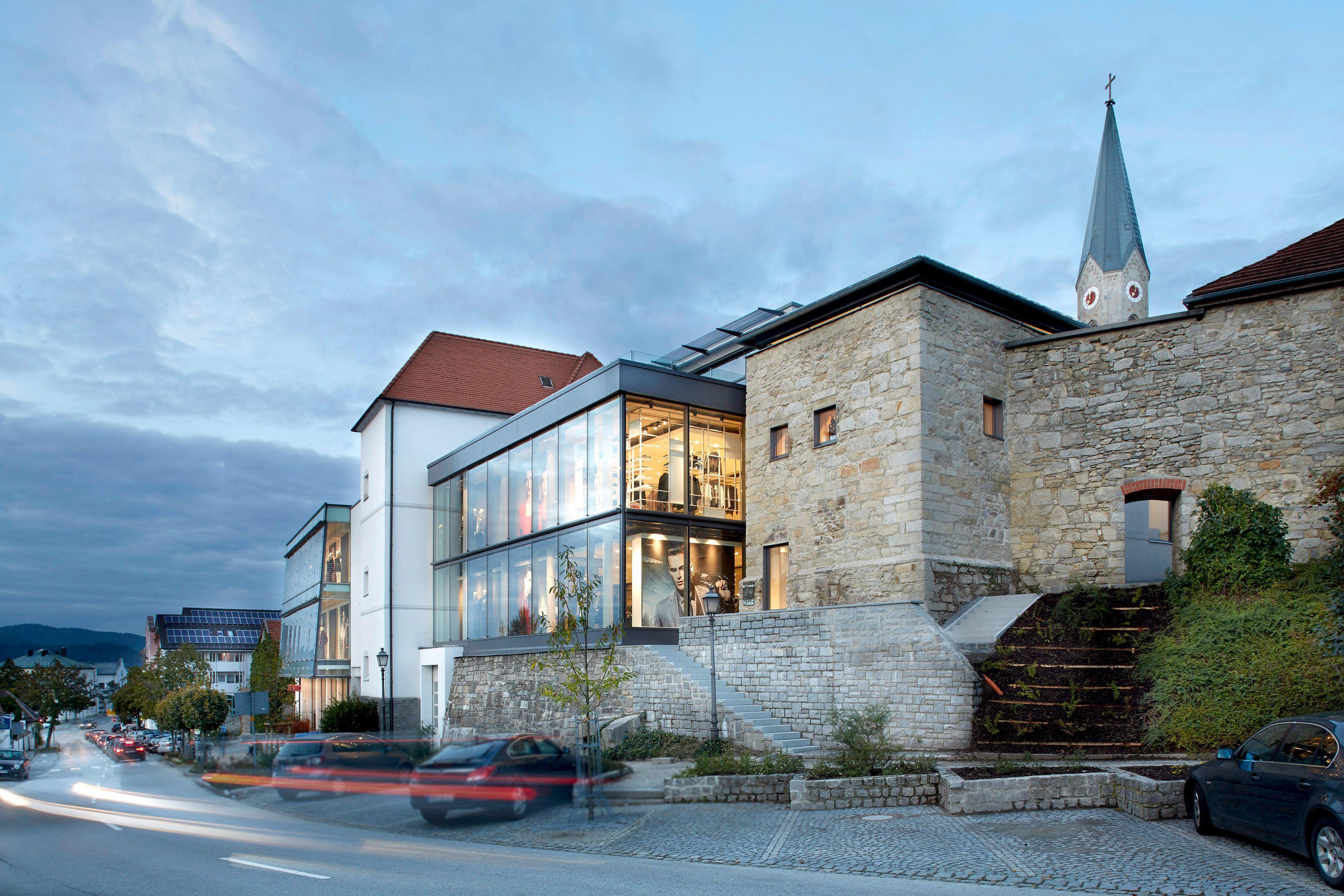 Modern glass building attached to historic stone structure with church spire at dusk.