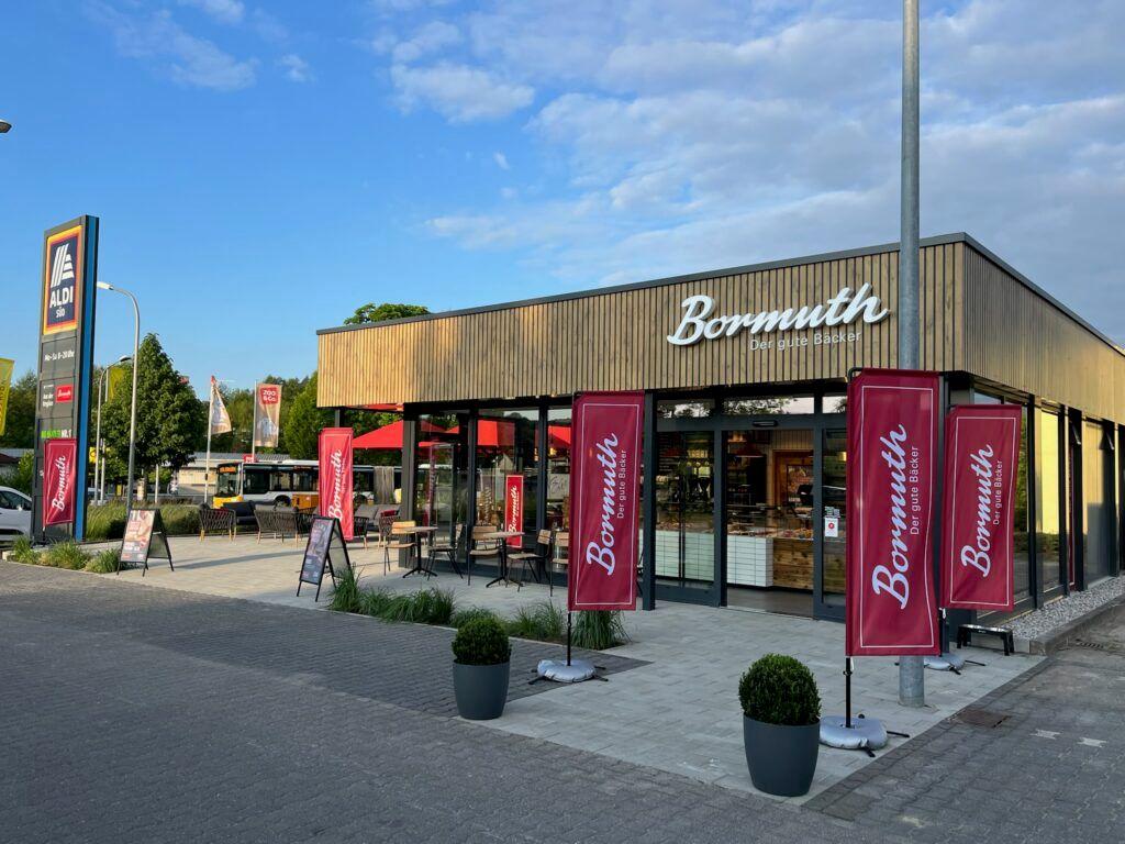 Modern Bormuth store with wooden facade and red banners under a partly cloudy sky.