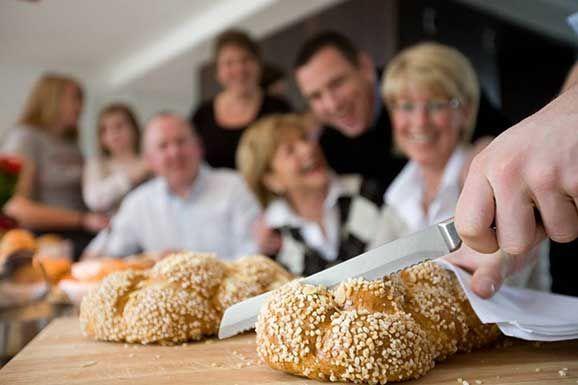 Hand cutting a sesame challah bread on a wooden board, with family in the background.