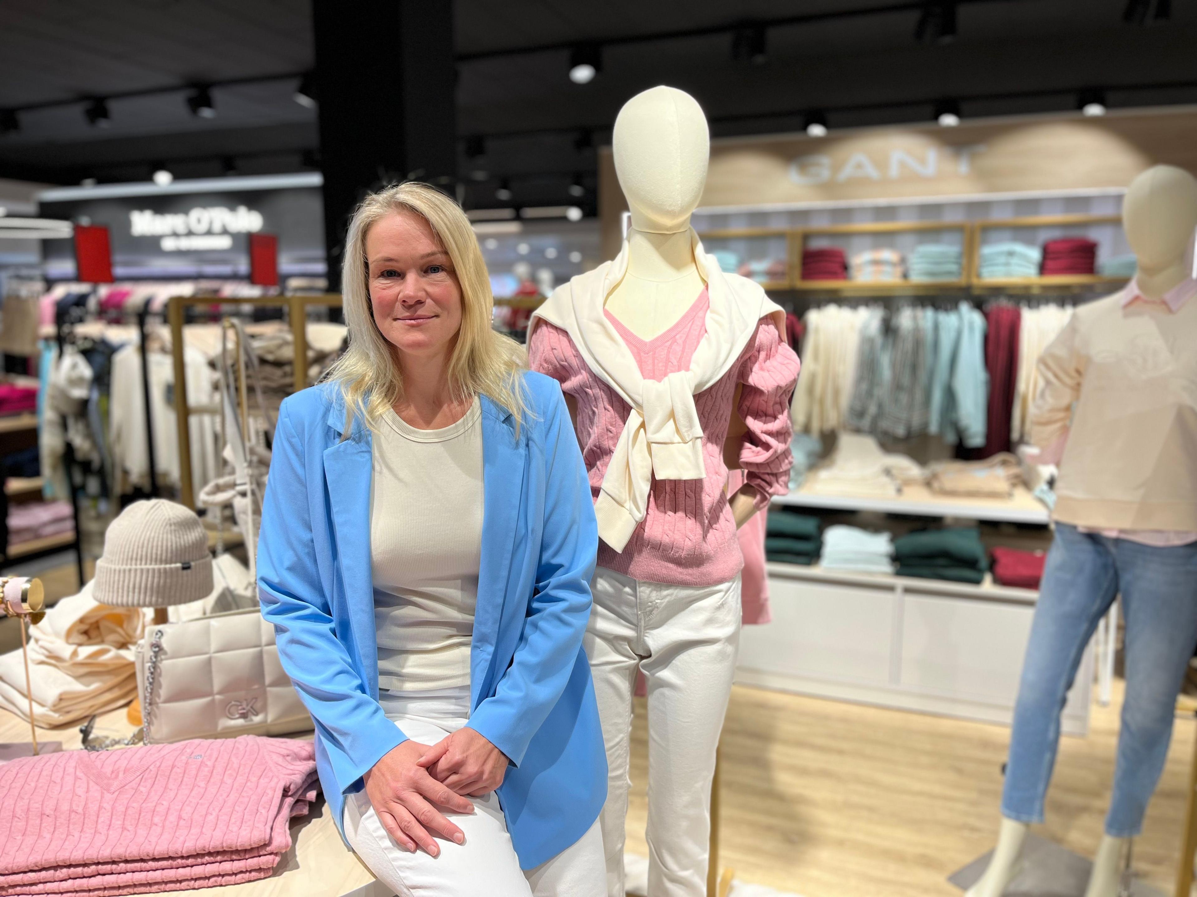 Woman in blue blazer sits beside mannequin with pink sweater in a clothing store.