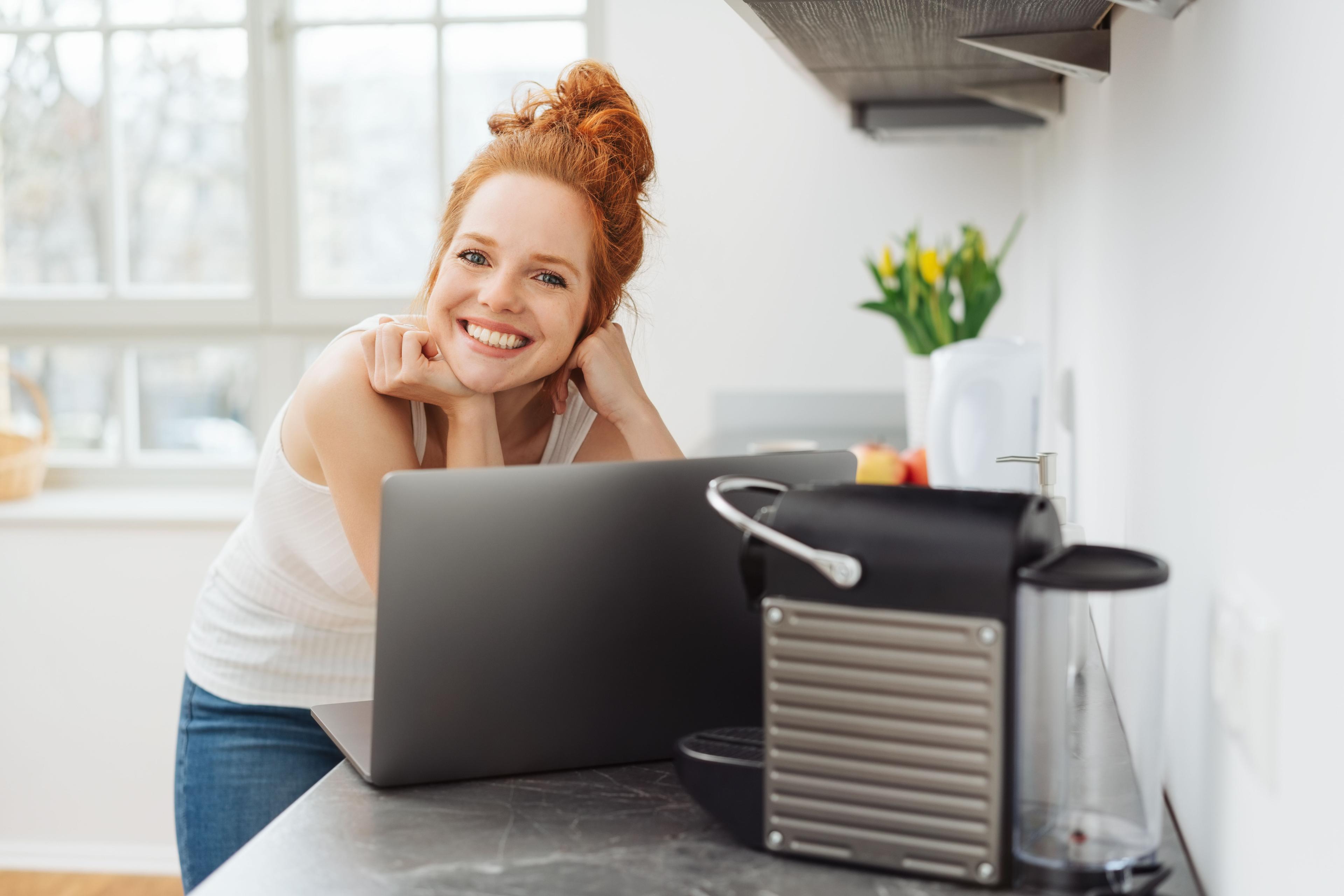 Happy redhead woman smiling at camera, working on laptop near a coffee machine.