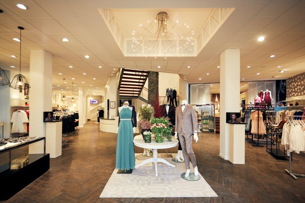 Brightly lit clothing boutique interior featuring mannequins displaying a dress and suit, and a staircase.