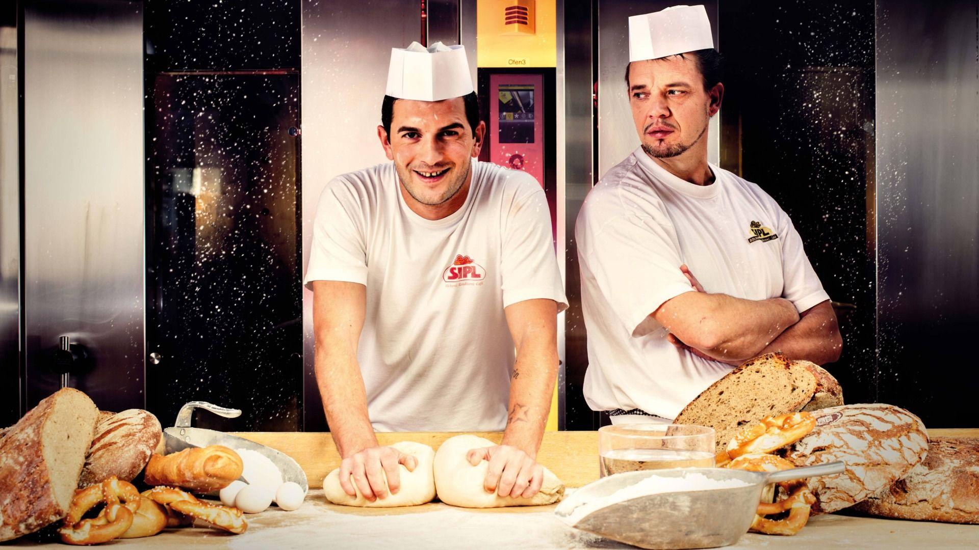 Two smiling bakers in white uniforms kneading dough among fresh loaves of bread.
